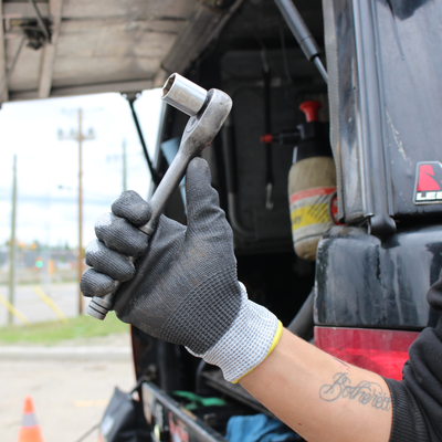 A close-up of a person's gloved hand holding a silver socket wrench in front of an open vehicle trunk, with a tattoo visible on the forearm.