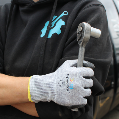 A close-up of a person wearing a black hoodie and a gray knit work glove with 'Spectra' branding, holding a metal socket wrench.